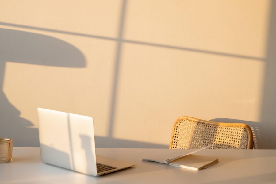 An empty, woven and wooden chair is pushed slightly away from a light wooden desk. There is both a laptop and a notebook open on the desk, but no one is in the frame. There is also light pouring into the office space through a large window.