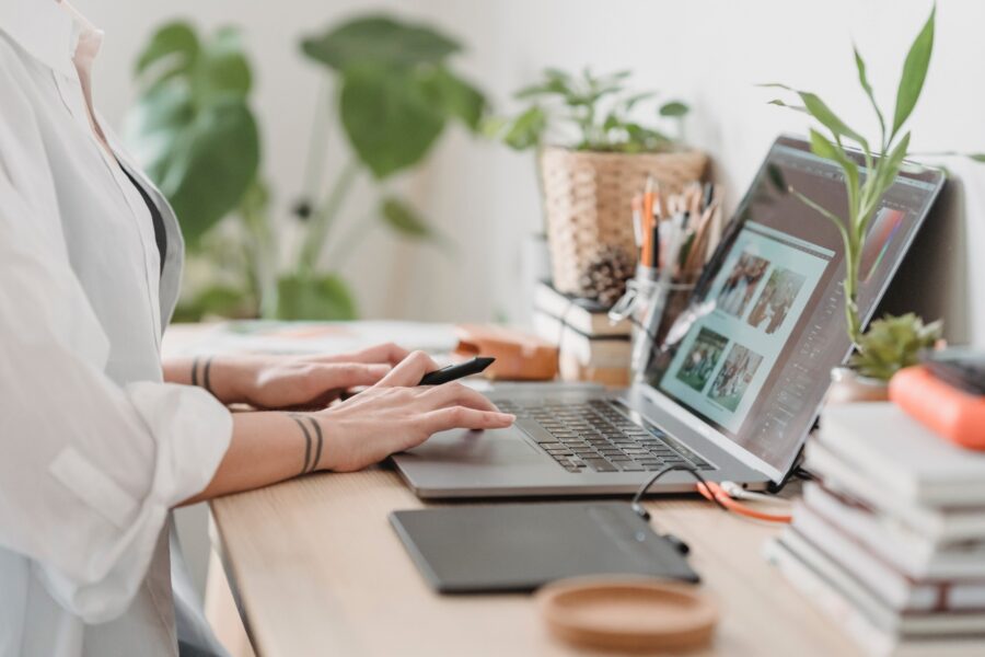 A white worker is sitting at a desk in their home office. They have a stylus in their hand and are using their laptop trackpad at the same time. Along with their laptop, there is also a drawing pad, books, office supplies like pens and paper, and plants on their desk. There is also a large plant in the background.