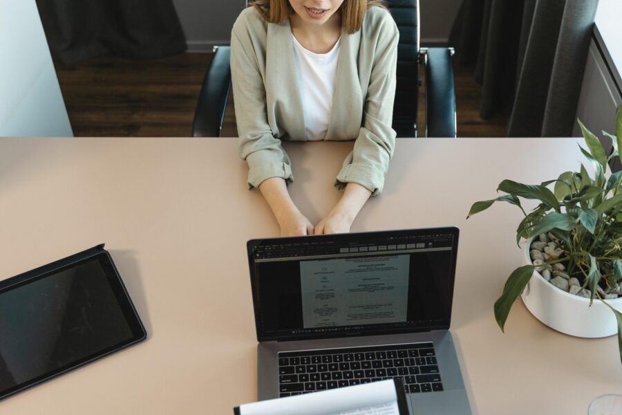 A woman is sitting at a large oak desk in a job interview. The person on the other side of the table is not in view—only their hands are, which are holding the interviewee's resume on a clipboard. There is also a laptop and tablet open on the desk facing the interviewer. The woman being interviewed looks calm and happy.