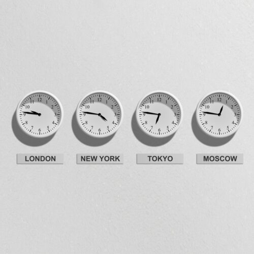 A white wall holds four white clocks in a row showing times in London, New York, Tokyo, and Moscow respectively. Below the clocks are white signs with black text that say the names of each city.