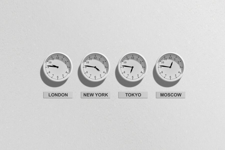 A white wall holds four white clocks in a row showing times in London, New York, Tokyo, and Moscow respectively. Below the clocks are white signs with black text that say the names of each city.