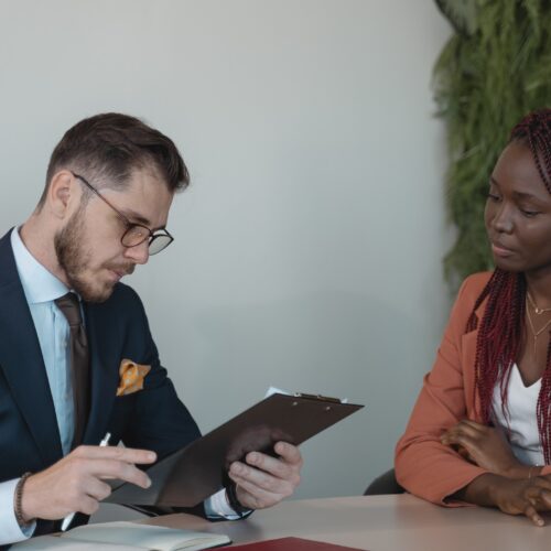 A man and woman are sitting at a wooden table in an office space. The man is looking at the woman's resume, which is clipped to a clipboard. The woman is watching the man review her resume, smiling gently. They are both in corporate workwear.