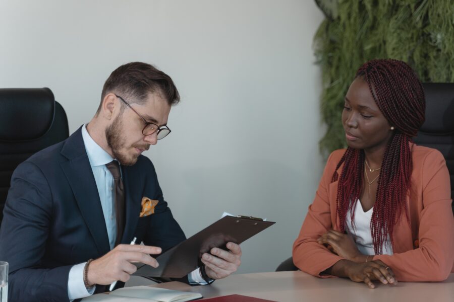 A man and woman are sitting at a wooden table in an office space. The man is looking at the woman's resume, which is clipped to a clipboard. The woman is watching the man review her resume, smiling gently. They are both in corporate workwear.