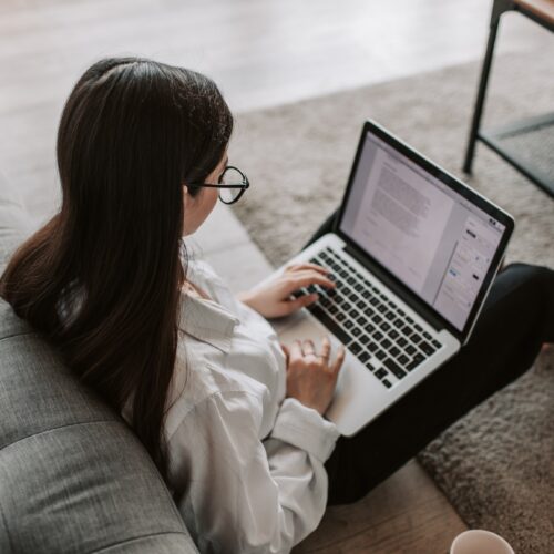 A young woman is sitting in her living room on the floor with her back against the sofa, typing on her laptop in her lap. She has a word-processing document open and is typing on the keyboard. She is wearing a white button-up shirt, black trousers, and large, black, thin glasses.