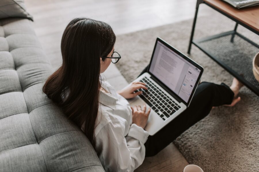 A young woman is sitting in her living room on the floor with her back against the sofa, typing on her laptop in her lap. She has a word-processing document open and is typing on the keyboard. She is wearing a white button-up shirt, black trousers, and large, black, thin glasses.