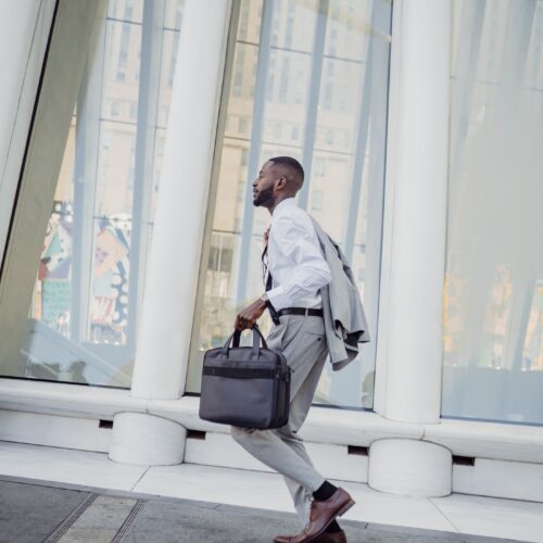 A man is running outside a sleek, white, modern building in grey trousers, a white shirt, and a grey matching blazer slung over his shoulder. He is also carrying a briefcase or laptop case, and has his head up, as if focused on his trajectory.