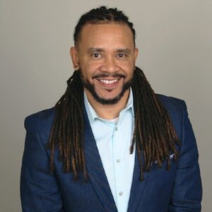 A headshot of Bradford Brown. He is smiling gently, looking at the camera. He has long, tidy dreadlocks pulled away from his face, a fresh looking, short beard, pierced ears, and a sharp navy blazer on over a light blue button-up shirt. 