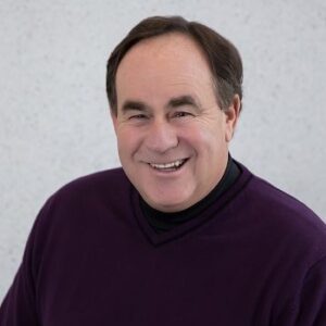 Stu Schlackman is a man with dark brown hair, thick eyebrows, and a gentle smile. He is wearing a dark knitted sweater in his headshot, which shows him smiling, looking into the camera, against a light grey background. 