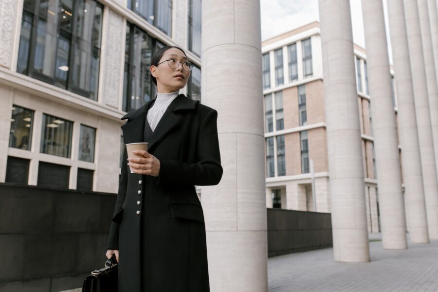 A woman dressed in corporate business clothing—a long, dark formal and fitted coat on top of a white blouse and blazer—is walking outside a commercial city space. She is carrying a coffee cup in one hand and a black briefcase in the other. She has her dark hair tied in a low ponytail and is wearing round, thin framed glasses. She is looking over her shoulder at a building nearby.