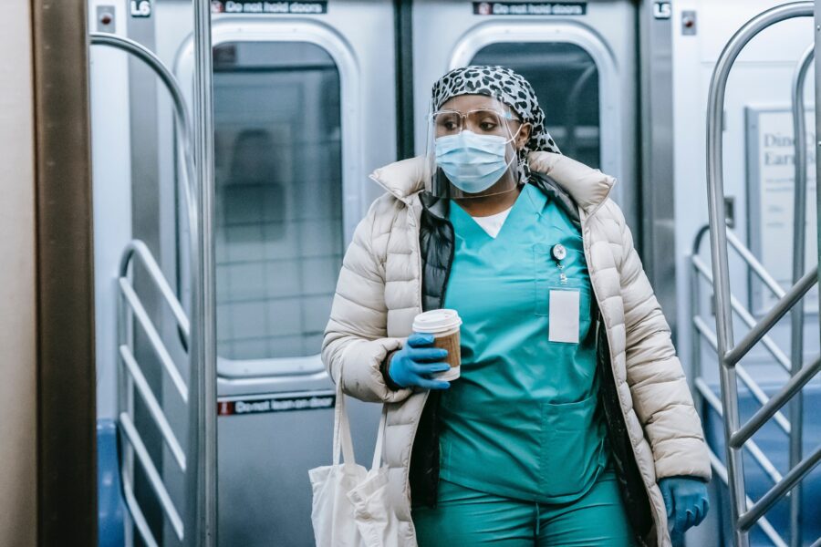 A Black woman is dressed in light teal medical scrubs, a winter parka, a grey leopard print scrub cap, a clear face shield, clear medical glasses, and medical gloves. She is holding a cup of coffee and a tote bag and is leaving a subway car at a station.