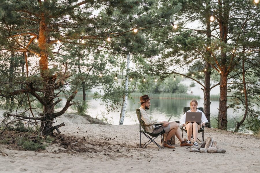 A man and woman are both sitting in folding lawn chairs in a wooded area. They are surrounded by a body of water, trees, and a campfire. They each have a laptop on their laps and are looking with concentration but content at their screens.