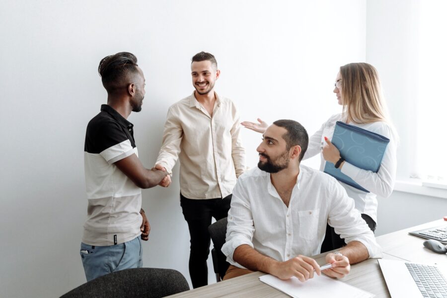 Three workers are introducing each other to a new employee. The new employee and two others are standing, shaking hands and making introductions. The third employee is sitting at a desk in front of them, turning back to say hello to the new employee. They are all dressed in business casual and are in a minimal office space with only a few papers and a computer nearby.
