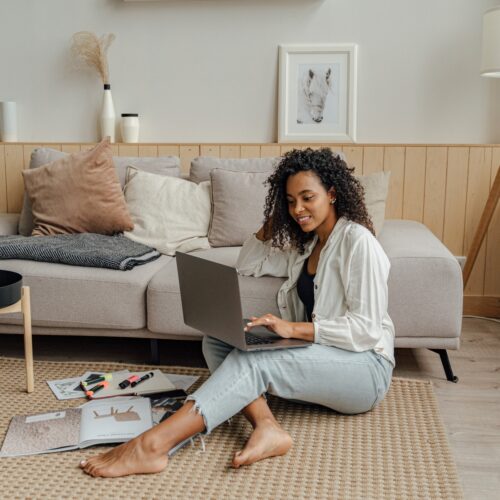 A Black woman sitting on the floor of her living room is working on her laptop. She has some paperwork and stationary items on the floor beside her. She is dressed in business casual clothing in neutral and pastel colours that match the tones of her boho interior design. She is smiling while working on her laptop.