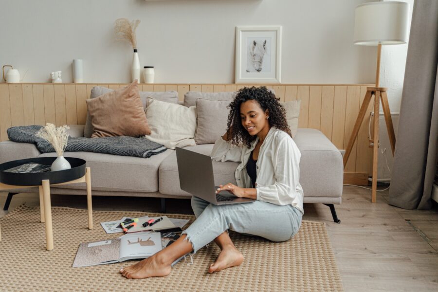 A Black woman sitting on the floor of her living room is working on her laptop. She has some paperwork and stationary items on the floor beside her. She is dressed in business casual clothing in neutral and pastel colours that match the tones of her boho interior design. She is smiling while working on her laptop.