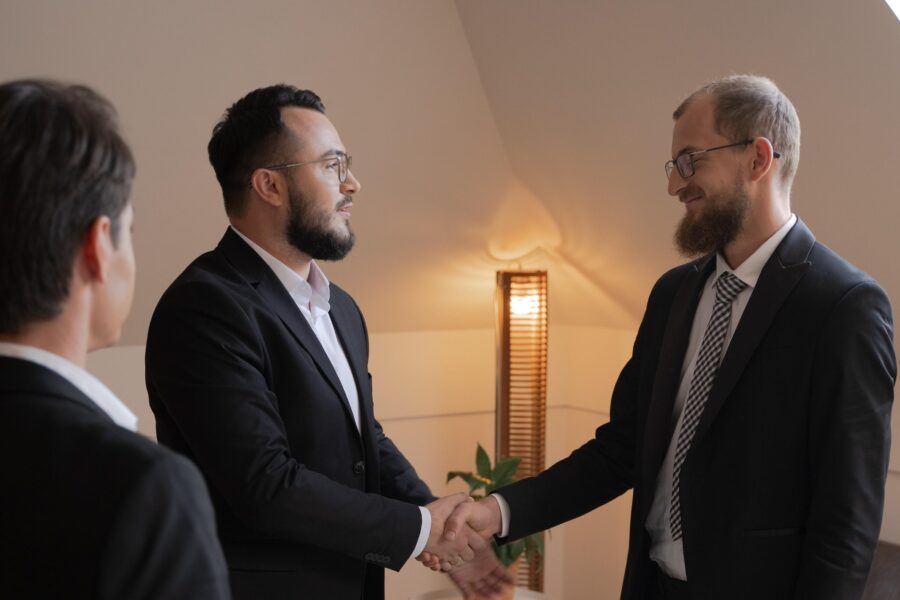Two businessmen are facing each other, shaking hands and smiling gently. There is one other person looking on at them shaking hands. They are all in a neutral, contemporary-looking office space with a modern, wooden, warm-toned lamp in the background and light, clean walls.