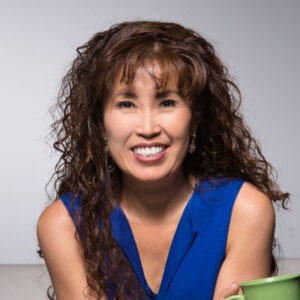 A headshot of Yoon Cannon. She is a Korean-American woman with long, light brown curly hair and a sleek, light fringe across her forehead. She has a brilliant smile and is posed leaning toward the camera invitingly here, wearing an electric blue blouse. 