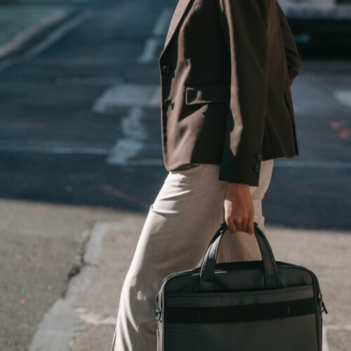 A woman wearing a brown blazer and khaki-coloured trousers is crossing a busy city street at a cross-walk, carrying a laptop case in one hand. The sun is low, shining on her at street-level, suggesting the end of a working day.